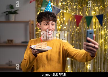 Ferne Gratulation. Porträt eines jungen Mannes, der sein Handy in der Hand hält, der der Webcam zuwinkt, einen Partyhut trägt und zu Hause seinen virtuellen Geburtstag feiert. Fernsteuerung Stockfoto