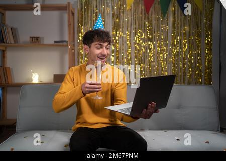 Ein Mann, der ein Videogespräch führt, feiert seinen Geburtstag online mit Freunden, die einen Laptop benutzen und ein Glas Champagner halten. Virtuelle Geburtstagsparty zu Hause. Stockfoto