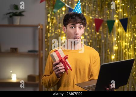 Zweifelnder Mann in Verwirrung feiert Geburtstag zu Hause mit Laptop und Geschenk und versteht nicht, was zu tun ist Stockfoto