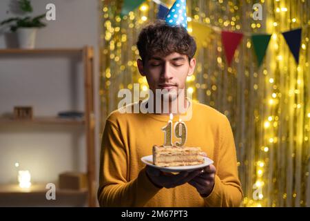 Ein junger Mann mit einem Konus-Partyhut, der sich seinen Geburtstagskuchen mit Kerzen Nummer 19 während der Feier zu Hause wünscht. 19. Geburtstag Stockfoto