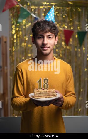 Zum 18. Geburtstag. Ein junger araber mit Partyhut, der Kuchen mit 18 Kerzen hält und zu Hause Geburtstag feiert und in die Kamera schaut Stockfoto