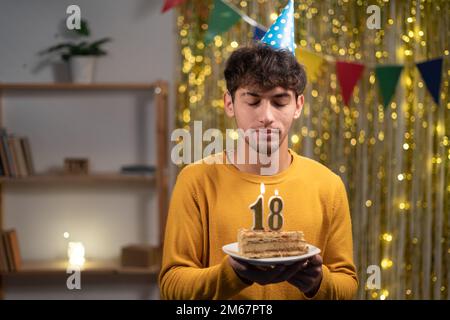 Ein junger Mann mit einem Konus-Partyhut, der sich seinen Geburtstagskuchen mit Kerzen Nummer 18 während der Feier zu Hause wünscht. Geburtstag der Achtzehnten Stockfoto