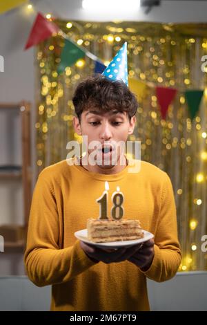 Arabische Schülerin in Cone-Party-Hut wünscht sich und bläst Kerzen Nummer 18 auf Geburtstagskuchen während der Feier zu Hause. Geburtstag der Achtzehnten. Stockfoto