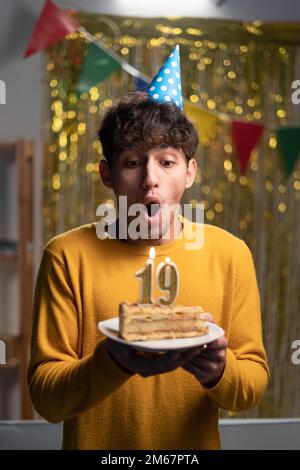 Arabische Schülerin in Cone-Party-Hut wünscht sich und bläst Kerzen Nummer 19 auf Geburtstagskuchen während der Feier zu Hause. 19. Geburtstag. Stockfoto