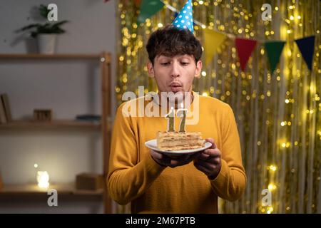 Arabische Schülerin in Cone-Party-Hut wünscht sich und bläst Kerzen Nummer 17 auf Geburtstagskuchen während der Feier zu Hause. Seventeens Geburtstag Stockfoto