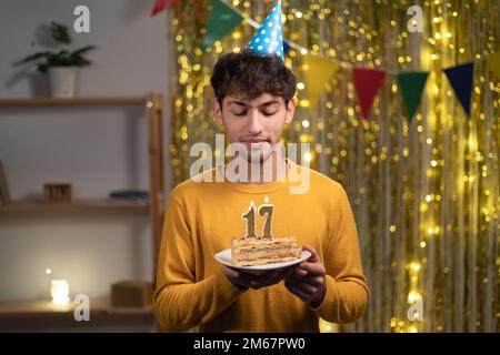 Ein junger Mann mit einem Konus-Partyhut, der sich seinen Geburtstagskuchen mit Kerzen Nummer 17 während der Feier zu Hause wünscht. Seventeens Geburtstag Stockfoto