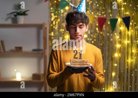 Ein Typ feiert zu Hause den 21. Geburtstag, hält einen Kuchen mit Kerzen und wünscht sich was Stockfoto