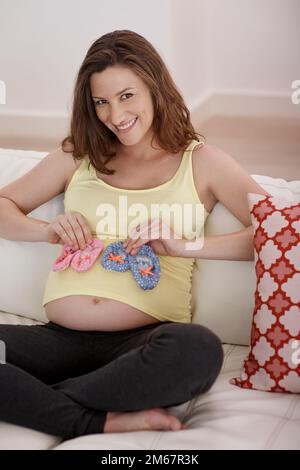 Pink oder Blau, beides reicht. Eine junge schwangere Frau, die ein Paar pinkfarbene und blaue Babyschuhe gegen ihren Bauch hält. Stockfoto