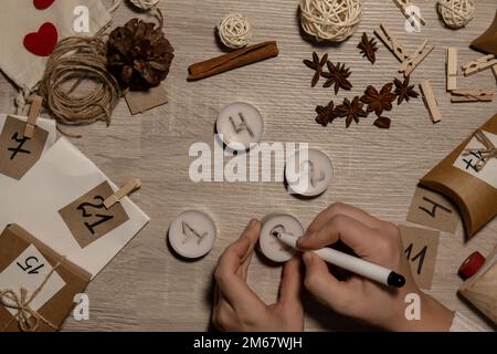 Unerkennbare junge Frau malt Zahlen auf Teekerzen. Frau macht hausgemachte Adventskalender mit eigenen Händen gemacht Schritt für Schritt selbstgemachtes Kunsthandwerk Machen Sie es selbst. Vorbereitung auf das weihnachtskonzept. Saisonale Aktivitäten für Kinder, Familienurlaub im Winter. Umweltfreundlich Geschenke. Öffnen Sie das Paket jeden Tag Stockfoto