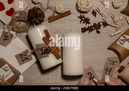 Eine nicht erkennbare junge Frau malt Zahlen auf Kerzen. Frau macht hausgemachte Adventskalender mit eigenen Händen gemacht Schritt für Schritt selbstgemachtes Kunsthandwerk Machen Sie es selbst. Vorbereitung auf das weihnachtskonzept. Saisonale Aktivitäten für Kinder, Familienurlaub im Winter. Umweltfreundlich Geschenke. Öffnen Sie das Paket jeden Tag Stockfoto