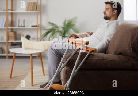 Junger Mann mit verletztem Bein, der zu Hause auf dem Sofa sitzt, in der Rehabilitation mit seinen Krücken Stockfoto