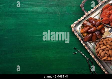 Gebetsperlen und ein Tablett mit Nüssen, Datteln, getrockneten Aprikosen auf einem dunkelgrünen Holztisch. Gesundes Essen für Breakfasts in Ramadan. Flach verlegt Stockfoto