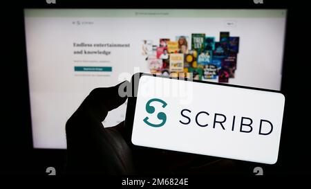 Person, die ein Mobiltelefon mit dem Logo des US-Verlagsunternehmens Scribd Inc. Auf dem Bildschirm vor der Unternehmenswebseite hält. Konzentrieren Sie sich auf das Display des Telefons. Stockfoto