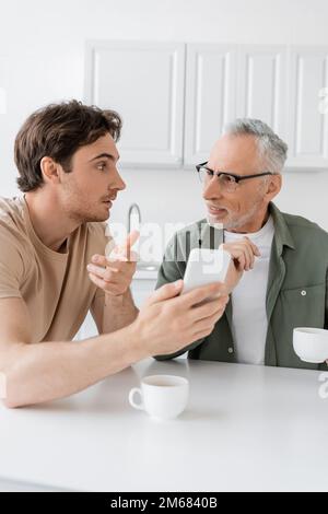 Der Mann zeigte nicht auf das Smartphone, während er mit einem lächelnden Vater in der Nähe von Kaffeetassen in der Küche sprach, Bild von Stock Stockfoto