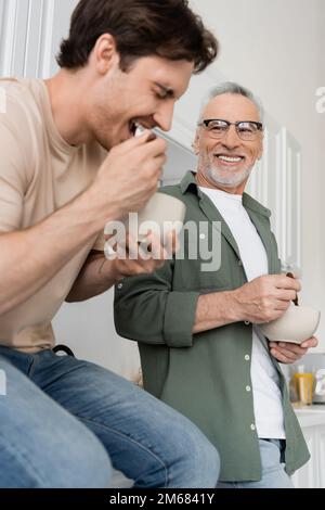 Ein glücklicher Mann mit Brille, der den jungen Sohn beim Frühstück ansieht und über ein verschwommenes Vordergrund- und Stockbild lacht Stockfoto