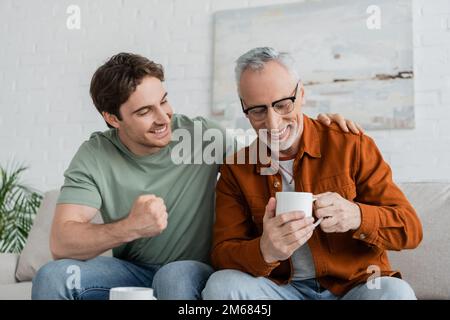 Aufgeregter Mann zeigt Gewinngeste, während er mit einem lächelnden Vater spricht, der zu Hause eine Teetasse auf dem Sofa hält, Stockbild Stockfoto