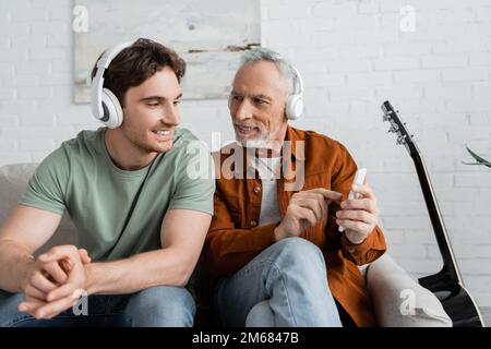 Grauer Mann mit kabellosen Kopfhörern, der sein Smartphone in der Nähe eines lächelnden Sohnes hält, Stockbild Stockfoto
