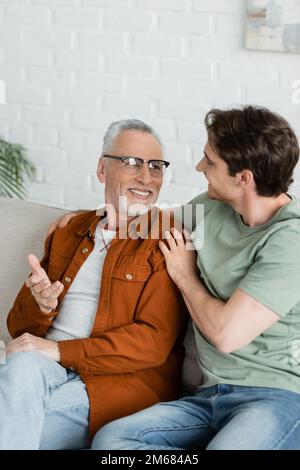 Glücklicher junger Mann, der lächelnd umarmt, und reifer Vater mit Brille, während er auf der Couch sitzt, Stockbild Stockfoto