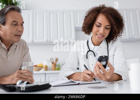 Lächelnder afroamerikanischer Arzt, der Glukometer in der Hand hält und in Notizbuch schreibt, Patient mittleren Alters, Stockbild Stockfoto