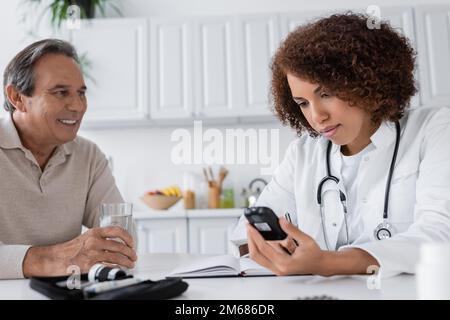 Lockiger afroamerikanischer Arzt, der Glukometer hält und in Notizbuch schreibt, fast lächelnder Patient mittleren Alters, Stockbild Stockfoto