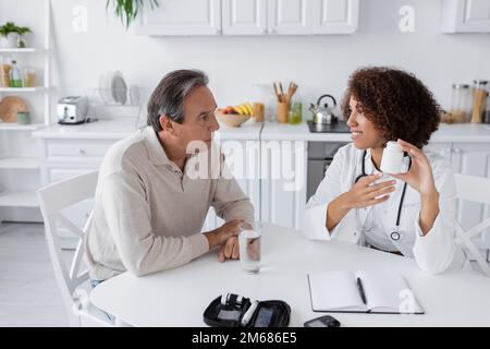 Lächelnder afroamerikanischer Arzt in weißem Mantel, Flasche mit Pillen in der Nähe von Patienten mittleren Alters mit Diabetes, Stock Image Stockfoto