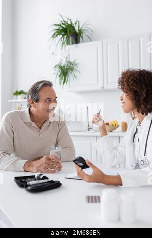 Lockiger afroamerikanischer Arzt, der das Glukometer in der Nähe eines lächelnden Patienten mittleren Alters hält, Stockbild Stockfoto