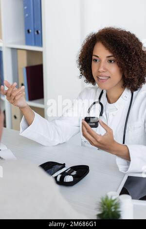 Fröhlicher afroamerikanischer Arzt mit Glukometer und Gespräch mit Patienten mittleren Alters mit Diabetes, Stock Image Stockfoto
