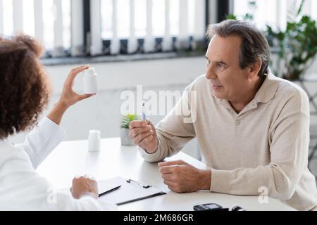 Lächelnder Patient hält Lanzettenstift in der Nähe eines afroamerikanischen Arztes mit Pillen in der Klinik, Stock Image Stockfoto