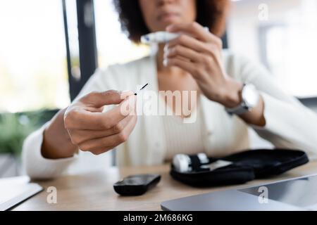 Zugeschnittene Ansicht einer verschwommenen afroamerikanischen Frau, die den Teststreifen des Glukometers im Büro hält, Stockbild Stockfoto