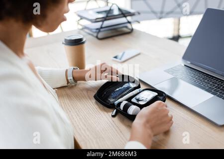 Freigestellte Ansicht einer afroamerikanischen Geschäftsfrau, die Diabetes-Kit in der Nähe von Geräten und Kaffee im Büro hält, Bild von Stock Stockfoto