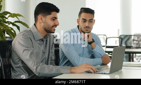 Zwei indische Mitarbeiter sitzen in einem modernen Büro mit einem Laptop, und junge Kollegen diskutieren über das Projekt. Arabische, seriöse, männliche Geschäftsmänner der Millennials zufrieden Stockfoto
