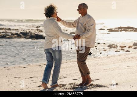 Glückliches älteres Paar, das lächelt und zusammen auf dem Strand tanzt. Reifes Paar, das sich am Meer amüsiert. Fröhliches Seniorenpaar genießt es Stockfoto