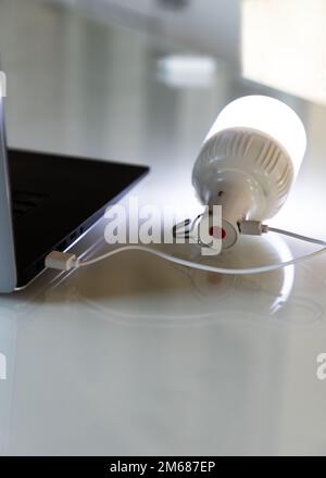 Die tragbare wiederaufladbare LED-Notleuchte mit integriertem Akku wird über ein USB-Kabel von einem Laptop aufgeladen. Lichtquelle bei Stromausfall. Vertikales Sho Stockfoto