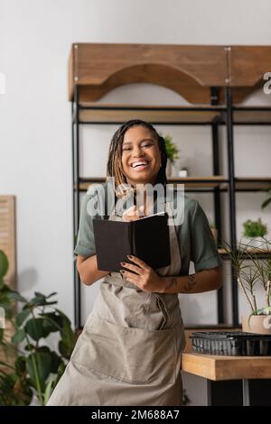 Lachender afroamerikanischer Florist mit Dreadlocks, der ein Copybook hält und in der Blumenhandlung auf die Kamera schaut, Aktienbild Stockfoto