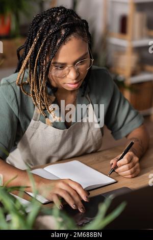 ein afroamerikanischer Florist mit Dreadlocks, der einen Laptop benutzt, während er einen Stift in der Nähe eines leeren Copybooks im Blumenladen hält, ein Aktienbild Stockfoto