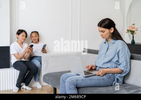 Mutter benutzt Laptop in der Nähe verschwommener asiatischer Kinder mit Handys zu Hause, Stock Image Stockfoto