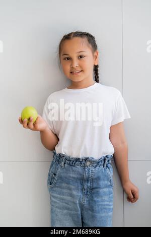 Lächelndes asiatisches Mädchen mit reifem Apfel und Blick auf die Kamera in der Nähe der Wand zu Hause, Stockbild Stockfoto