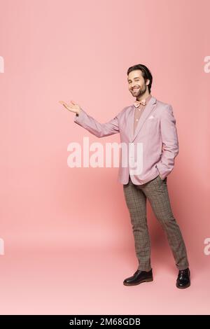 Volle Länge des positiven Hosts des Ereignisses, der mit der Hand auf einen rosafarbenen Hintergrund zeigt, Aktienbild Stockfoto