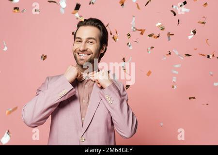 Lächelnder Veranstaltungsort in der Jacke mit Schleife unter Konfetti auf pinkfarbenem Hintergrund, Stockbild Stockfoto