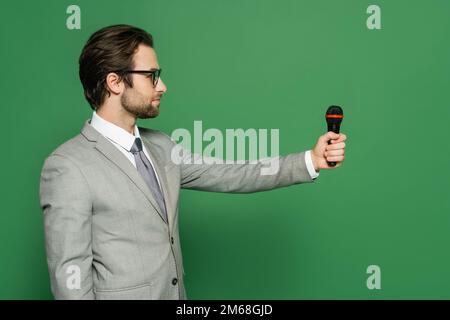 Seitenansicht des Nachrichtensenders im Anzug mit Mikrofon und Blick weg auf grünen Hintergrund, Stockbild Stockfoto