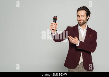 Fröhlicher Veranstaltungsort in formeller Kleidung, der auf das Mikrofon zeigt, isoliert auf grauem Stockbild Stockfoto