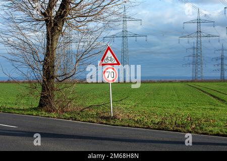 Bergheim, Deutschland. 29. Dezember 2022. Auf einer Landstraße steht ein Verkehrsschild mit einem Hinweis auf das Tempolimit von 70 km/h und „Achtung, Radfahrer“. Dahinter befinden sich zahlreiche Strommasten auf Feldern. Kredit: Viola Lopes/dpa/Alamy Live News Stockfoto