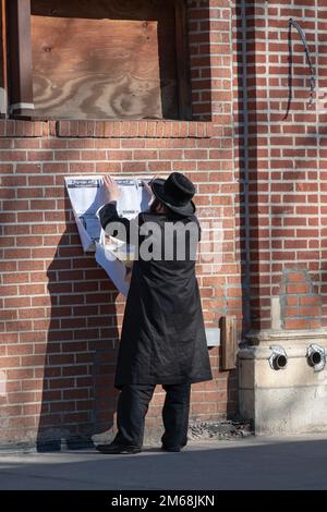 Ein chassidischer Jude hängt ein Schild mit einem Zeitplan für religiöse Ereignisse. Vor einer Synagoge auf der Lee Avenue in Williamsburg, Brooklyn, New York. Stockfoto