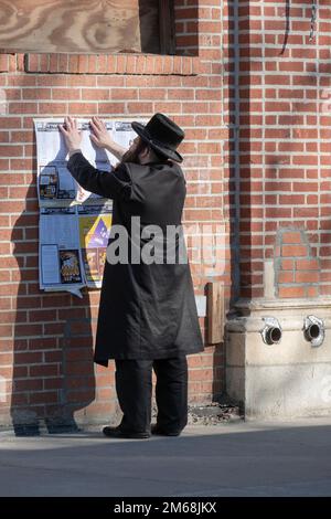 Ein chassidischer Jude hängt ein Schild mit einem Zeitplan für religiöse Ereignisse. Auf der Lee Avenue in Williamsburg, Brooklyn, New York. Stockfoto