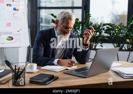 Geschäftsmann mittleren Alters, der auf einem Notebook in der Nähe eines Laptops schreibt und Kaffee im Büro macht, Bild von Stock Stockfoto