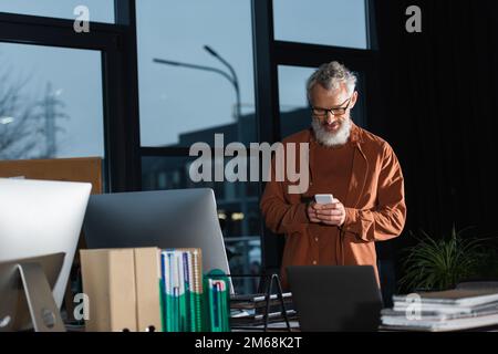 Grauhaariger Geschäftsmann, der ein Smartphone benutzt, während er am Arbeitsplatz neben Computern im Büro steht, Stockbild Stockfoto