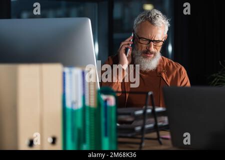 Ein gravierender, grauhaariger Geschäftsmann, der sich auf einem Smartphone in der Nähe von Computern und verschwommenen Ordnern im Büro unterhält, Aktienbild Stockfoto