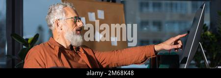 Seitenansicht eines bärtigen Geschäftsmanns, der während der Arbeit im Büro auf den Computerbildschirm zeigt, Banner, Aktienbild Stockfoto