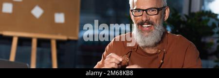 Ein glücklicher, bärtiger Geschäftsmann mit Brille, der Bitcoins in der Hand hält und auf Kamera, Banner, Stock-Bild schaut Stockfoto