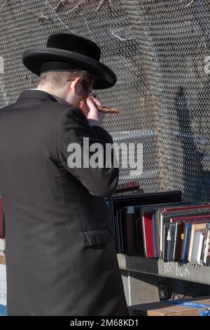 Ein orthodoxer jüdischer Mann mit rötlich-braunem Haar dreht sein langes Peyot, während er in einem Pop-up-Laden im Freien Bücher liest. In Brooklyn, New York. Stockfoto